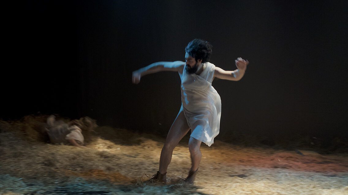 Tremor Teatro - Man, black hair and beard, running on a floor full of straw. Transparent white clothing.